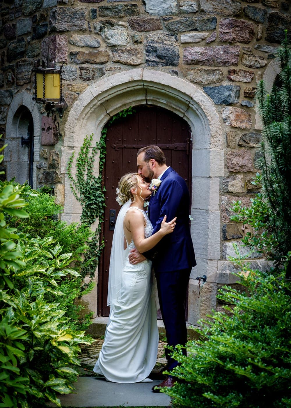 A bride and groom kissing in front of an arched wooden door set in a stone wall