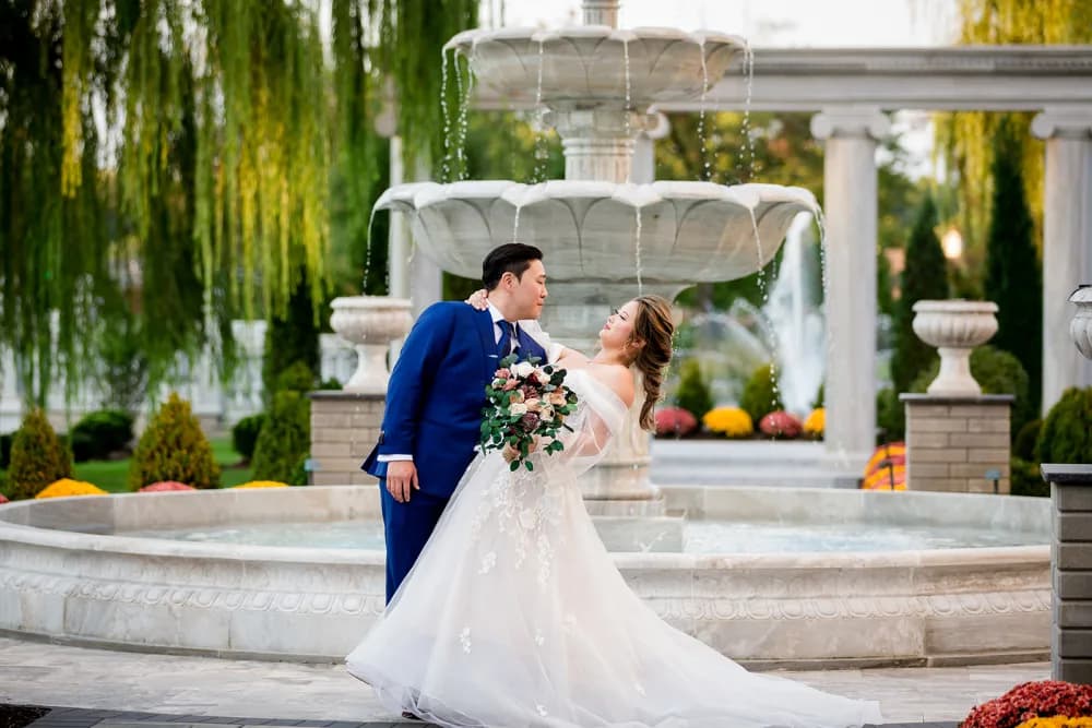 A bride and groom kissing beside a stone fountain beneath willow trees