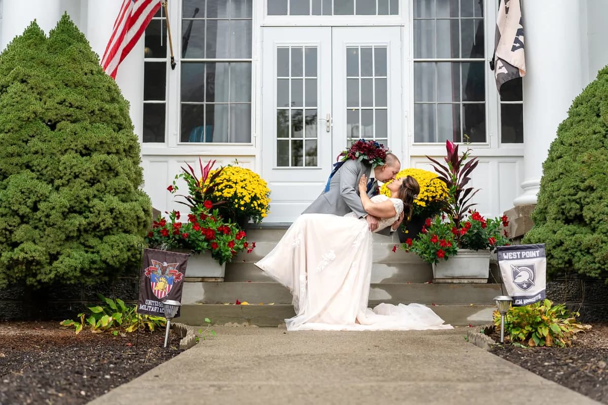 Bride and groom kissing in front of the white clubhouse and fall flowers at The Club at Picatinny