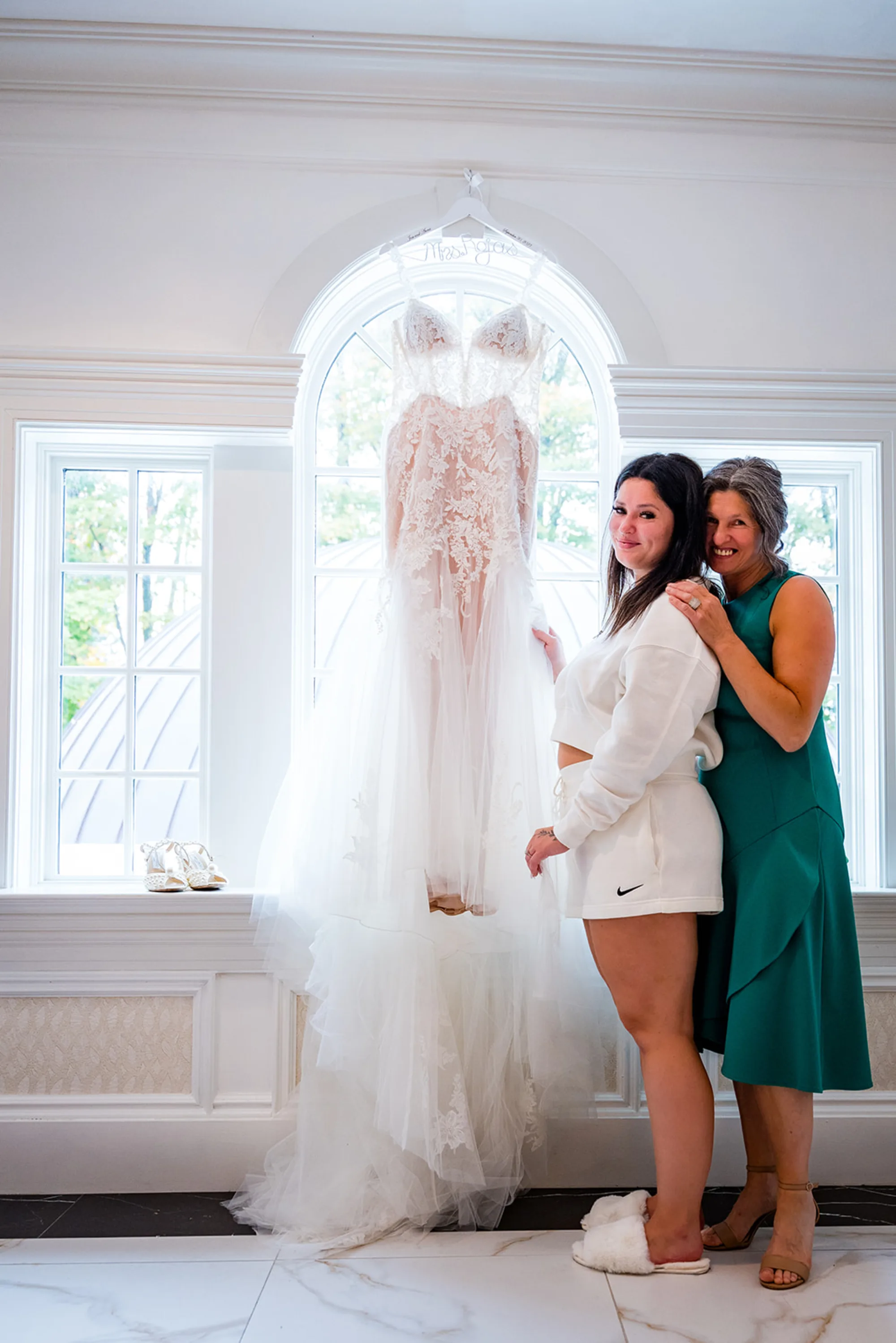 Anna with her mother beside the hanging wedding dress in a bright window at The Meadow Wood